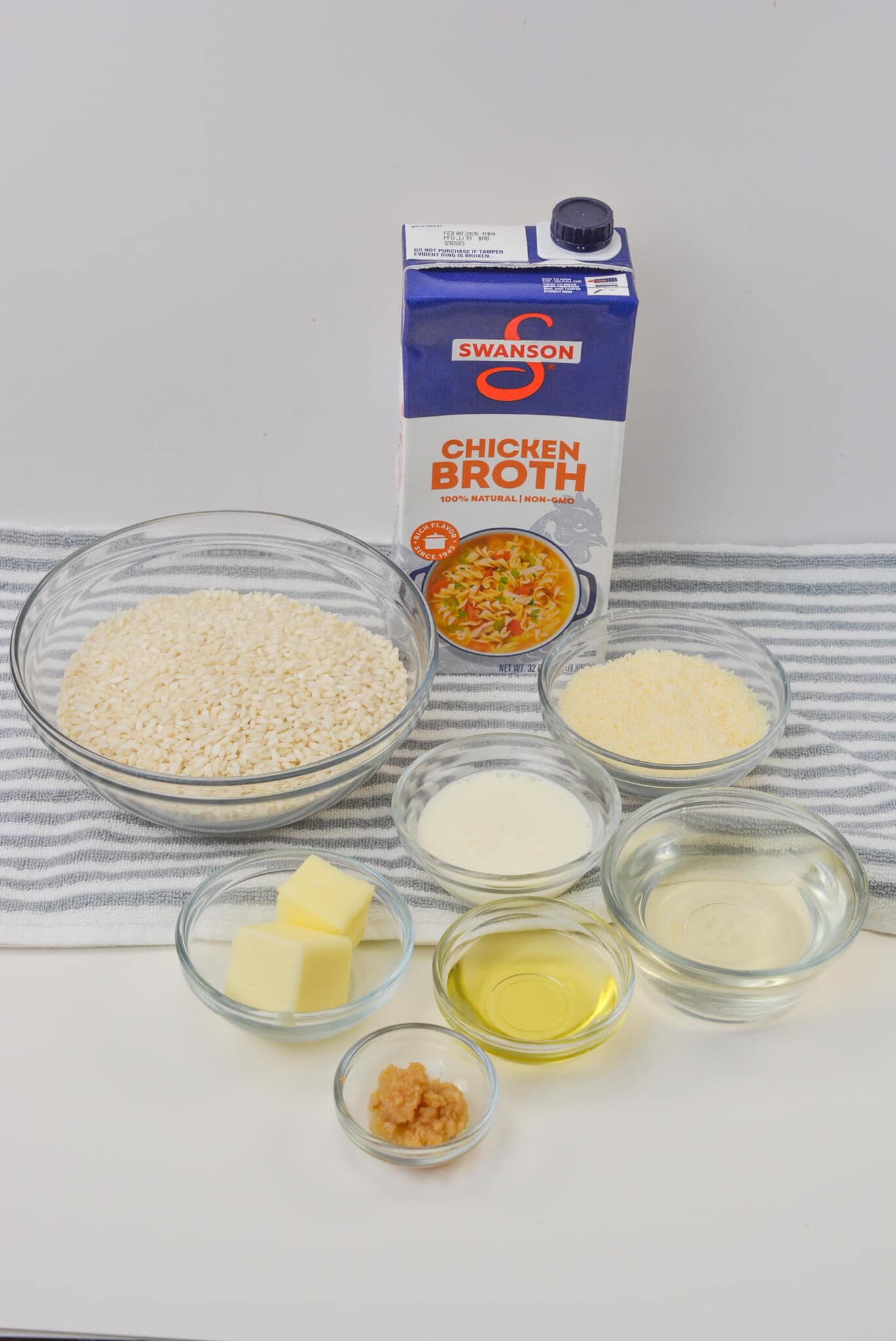 A delightful creamy risotto awaits, with ingredients elegantly arranged on a striped cloth: chicken broth, rice, cream, grated cheese, butter, olive oil, garlic, and white wine.
