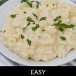 A plate of luscious, creamy risotto is garnished with chopped herbs, with utensils and fresh greens elegantly scattered in the background. Text at the bottom reads "Easy Creamy Risotto.