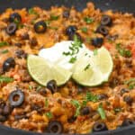 A meal brims with ground meat, black beans, olives, rice, and cheese. It's perfectly garnished with sour cream, lime slices, and chopped herbs.