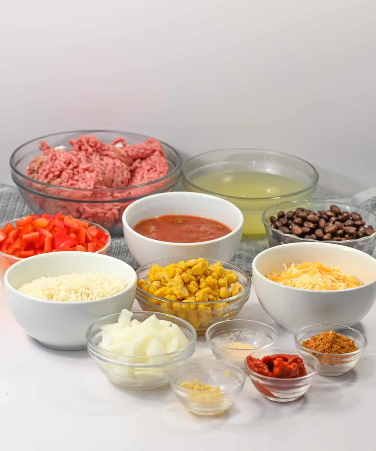 Various ingredients in bowls including ground meat, diced tomatoes, chicken broth, and black beans are arranged on a white surface, ready to create a delicious Mexican Beef and Rice Skillet with bell peppers, cheese, corn, onions, and spices.