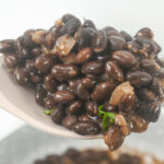 A spoonful of glossy, cooked beans lifted above a bowl, with beans visibly topped with minced onions and herbs.