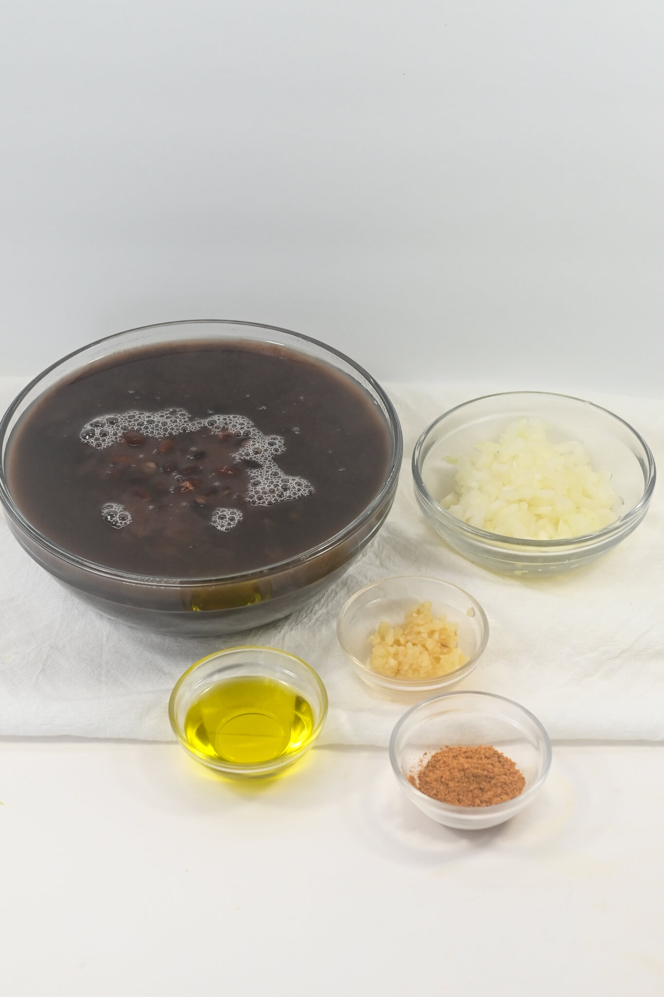 Bowl of Mexican black bean soup with small bowls of chopped onions, minced garlic, olive oil, and ground pepper arranged on a white surface.