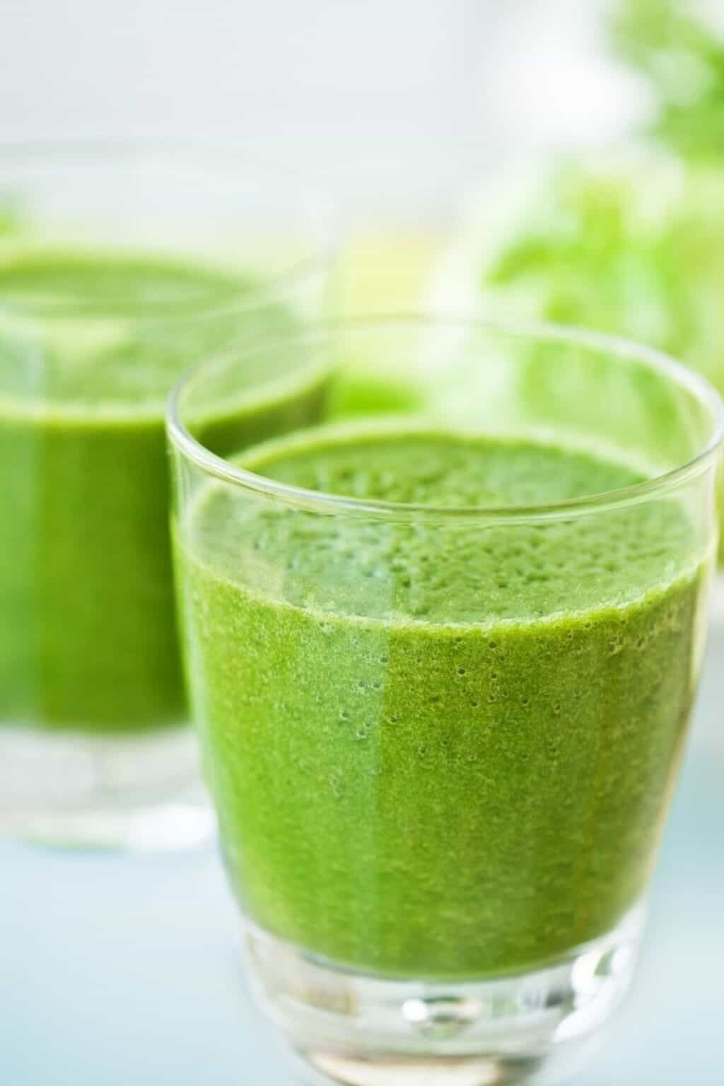 Two clear glasses filled with thick Green Goddess Smoothie, with a leafy green vegetable blurred in the background.