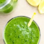 A glass of Green Goddess Smoothie with a yellow-striped straw sits next to lemon wedges and a jar, viewed from above.