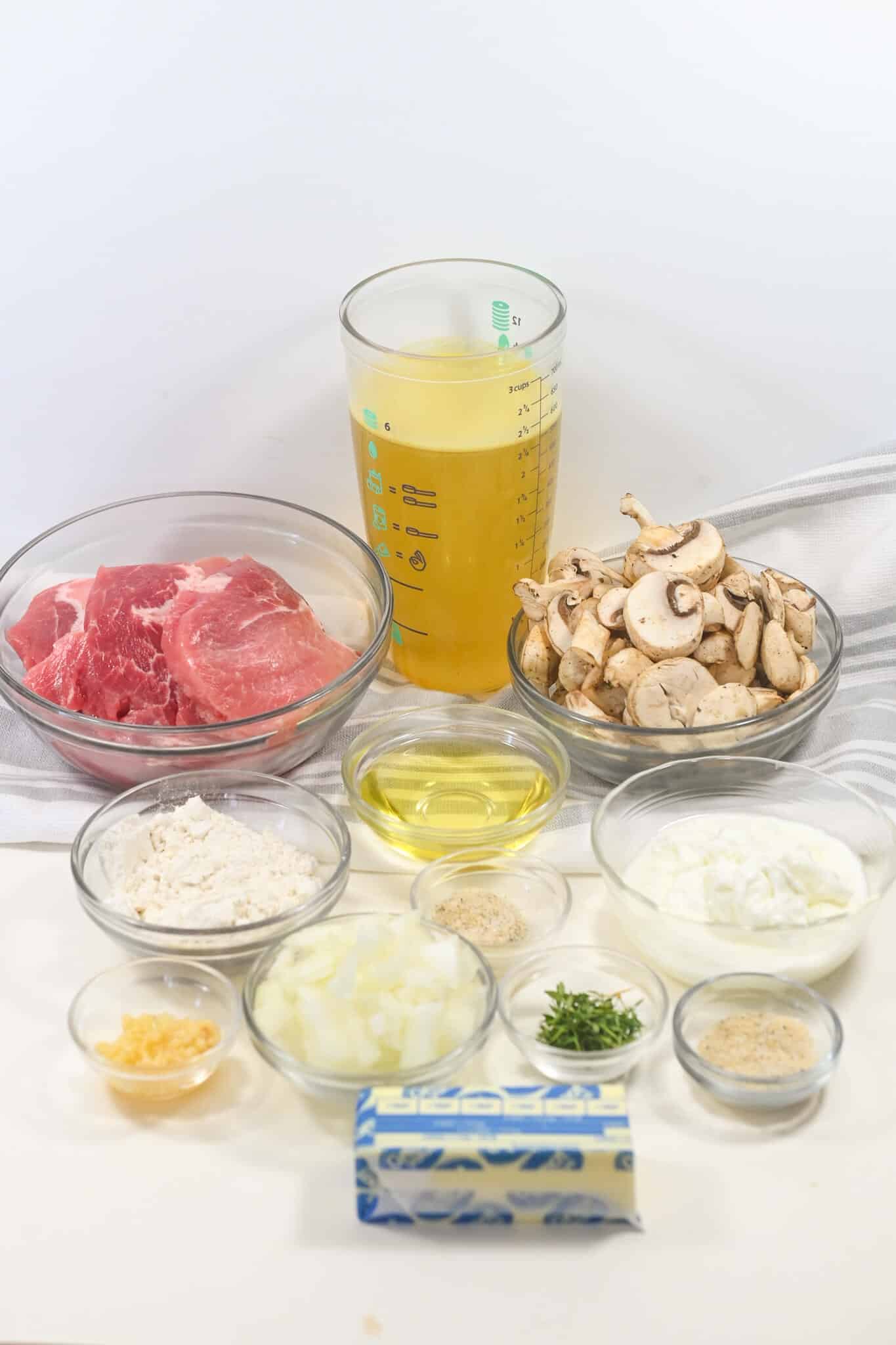 Ingredients for a recipe laid out, including pork chops, sliced mushrooms, flour, oil, onions, herbs, and broth.