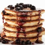 A stack of five pancakes topped with fresh blueberries and a drizzle of blueberry syrup on a white plate against a pristine white background.