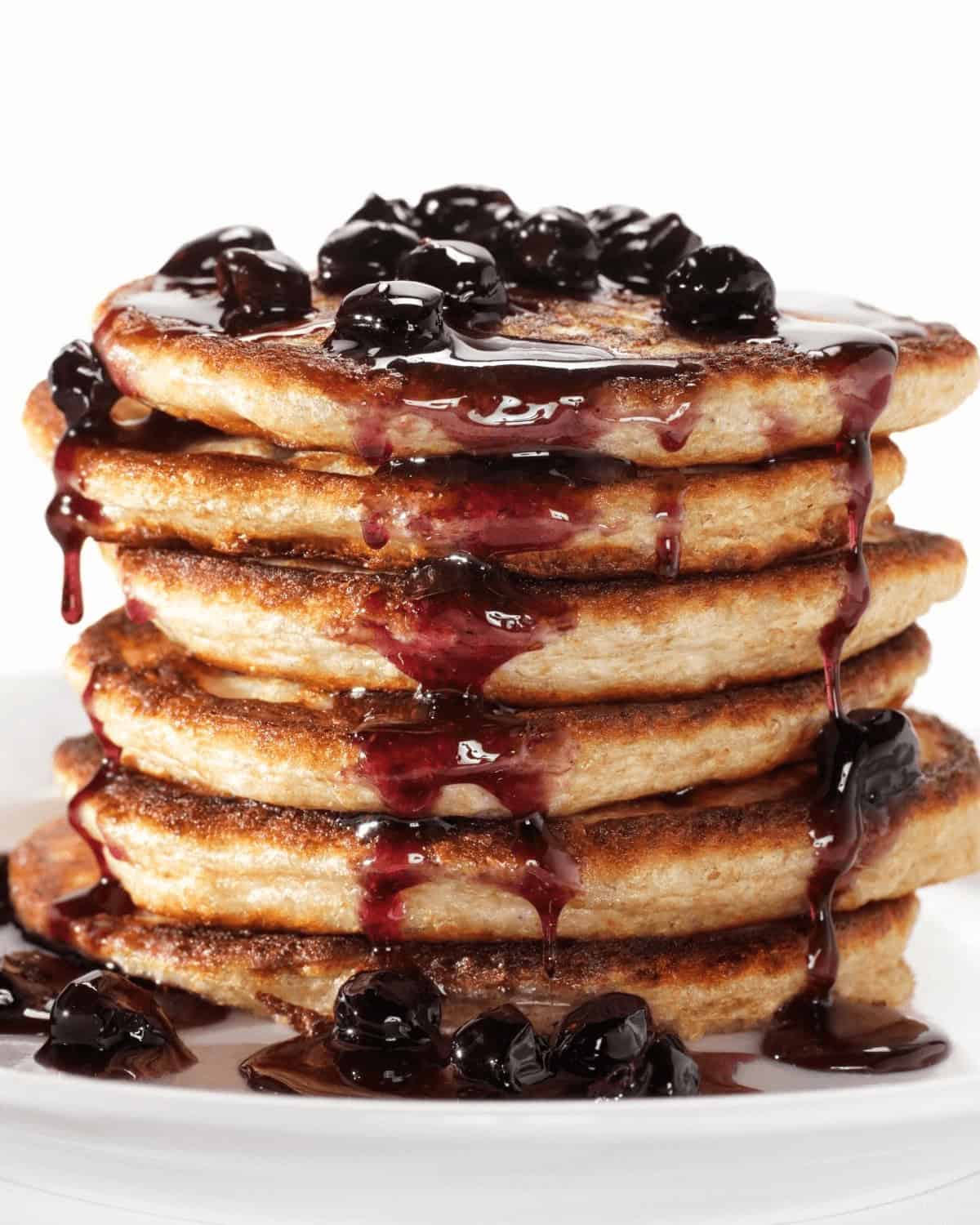 A stack of five pancakes topped with fresh blueberries and a drizzle of blueberry syrup on a white plate against a pristine white background.