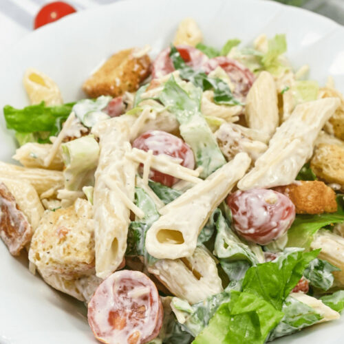 A plate of Caesar pasta salad, featuring penne noodles, halved cherry tomatoes, Romaine lettuce, and croutons, topped with shredded cheese and creamy Caesar dressing.