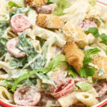 A bowl of Caesar pasta salad with cherry tomatoes, croutons, and chopped romaine lettuce, garnished with fresh basil.
