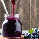 A jar of rich blueberry syrup being poured with a ladle, surrounded by fresh blueberries on a rustic wooden surface.