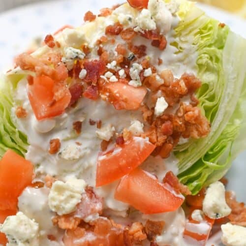 A classic Iceberg Wedge Salad, this dish features a crisp wedge of iceberg lettuce topped with diced tomatoes, bacon bits, blue cheese crumbles, and creamy dressing on a white plate.