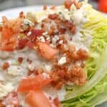 Enjoy our classic Wedge Salad: a crisp wedge of iceberg lettuce topped with creamy blue cheese dressing, diced tomatoes, savory bacon bits, and blue cheese crumbles—all beautifully arranged on a white plate.