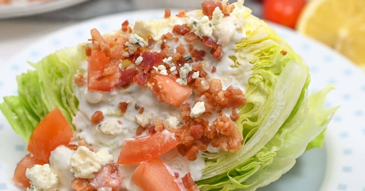 Enjoy our classic Wedge Salad: a crisp wedge of iceberg lettuce topped with creamy blue cheese dressing, diced tomatoes, savory bacon bits, and blue cheese crumbles—all beautifully arranged on a white plate.