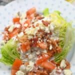 An meal features a crisp wedge of iceberg lettuce topped with diced tomatoes, bacon bits, blue cheese crumbles, and creamy dressing, all beautifully arranged on a white plate.