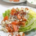 Enjoy a classic iceberg wedge salad— a crisp wedge of iceberg lettuce topped with diced tomatoes, blue cheese crumbles, savory bacon bits, and creamy dressing, all beautifully served on a white plate.