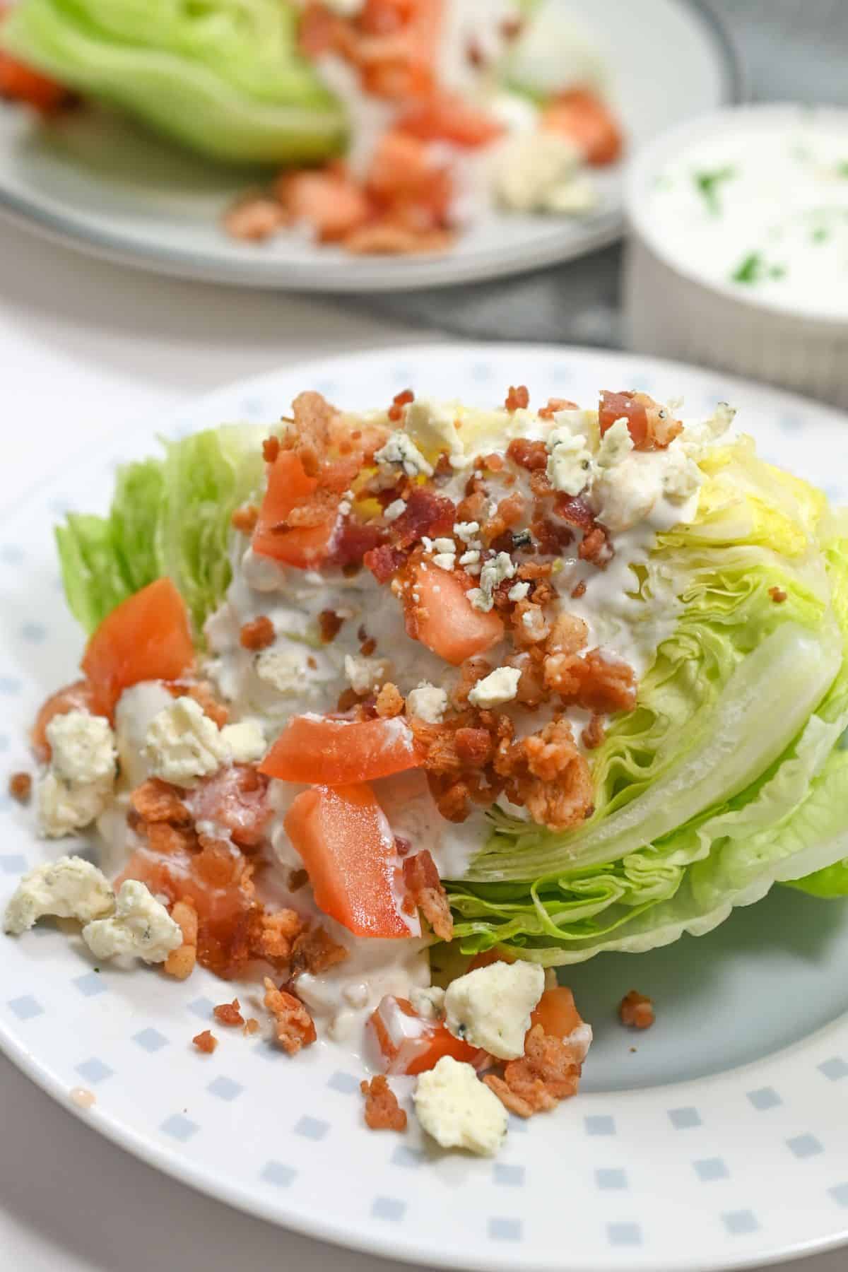 Enjoy a classic iceberg wedge salad— a crisp wedge of iceberg lettuce topped with diced tomatoes, blue cheese crumbles, savory bacon bits, and creamy dressing, all beautifully served on a white plate.