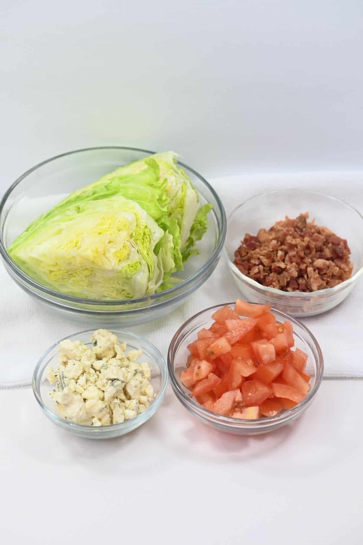 Four clear bowls containing iceberg lettuce wedges, chopped bacon, diced tomatoes, and crumbled blue cheese are arranged on a white surface with a white background, perfectly showcasing the classic ingredients for an Iceberg Wedge Salad.