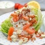 A classic Wedge Salad featuring crisp iceberg lettuce, topped with chopped tomatoes, crumbled bacon, blue cheese, and creamy dressing on a white plate.