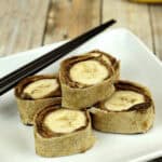Slices of rolled tortilla with embedded banana pieces on a white plate, resembling delightful\accompanied by black chopsticks.