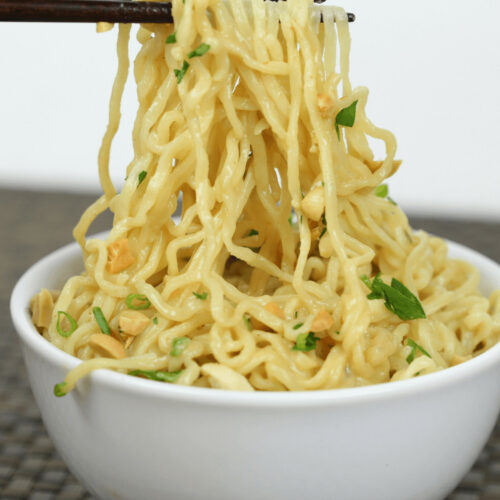 Chopsticks lifting spicy peanut noodles from a white bowl garnished with green onions and herbs, set on a woven mat.
