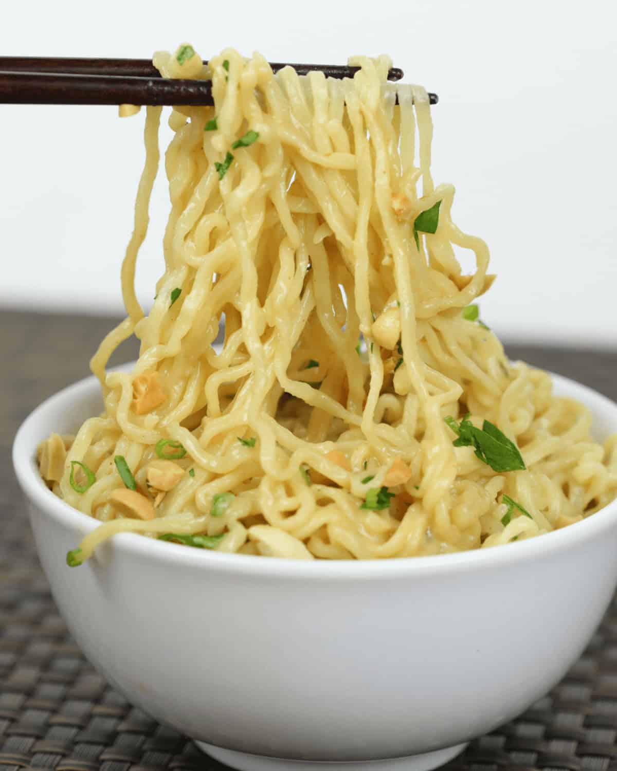 Chopsticks lifting spicy peanut noodles from a white bowl garnished with green onions and herbs, set on a woven mat.