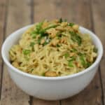 A bowl of cooked spicy instant noodles garnished with chopped herbs and crushed peanuts, displayed on a wooden surface.