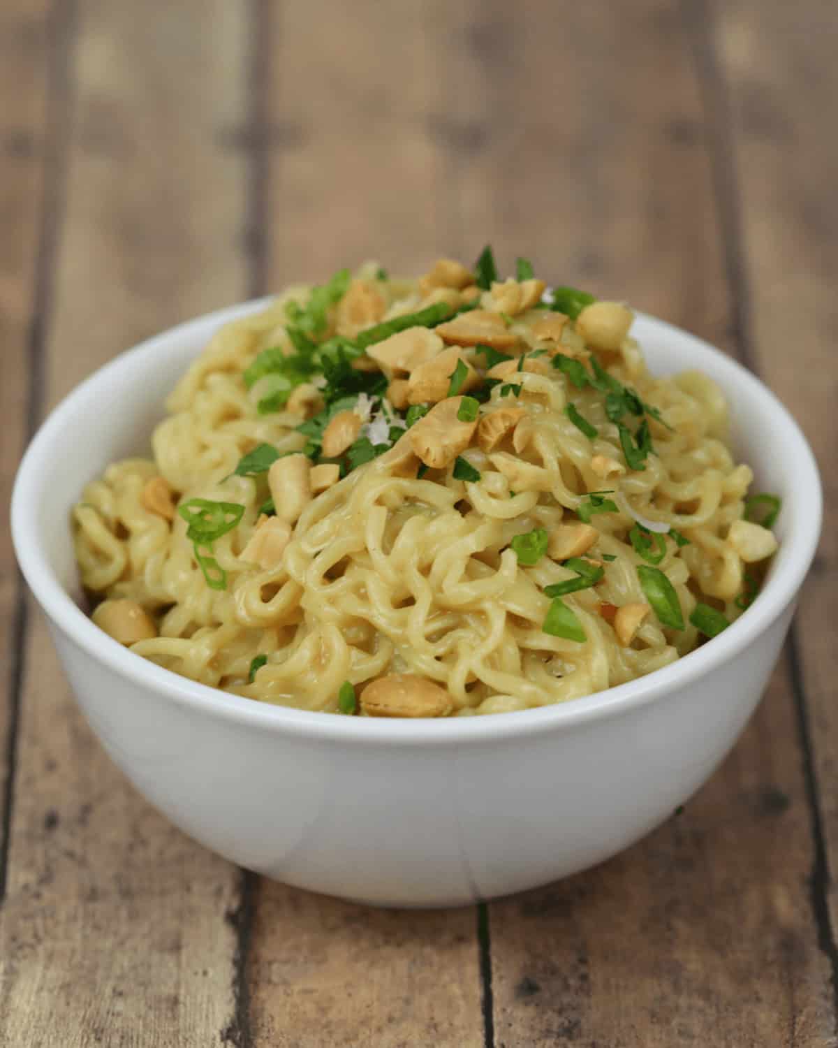 A bowl of cooked spicy instant noodles garnished with chopped herbs and crushed peanuts, displayed on a wooden surface.