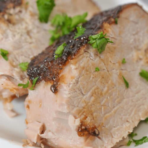 Tender slices of pork with crispy skin, beautifully garnished with fresh parsley on a white plate, evoke the rich flavors of a slow-cooked dish reminiscent of pork tenderloin in a crock pot.