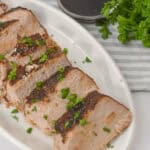 Succulent sliced roast pork tenderloin, slow-cooked to perfection in a crock pot, garnished with parsley on a white plate. It sits beside a small bowl of sauce and a bundle of fresh parsley for an elegant touch.