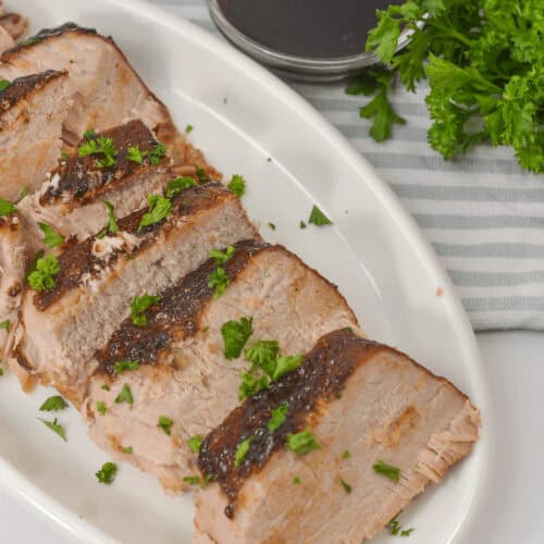 Succulent sliced roast pork tenderloin, slow-cooked to perfection in a crock pot, garnished with parsley on a white plate. It sits beside a small bowl of sauce and a bundle of fresh parsley for an elegant touch.