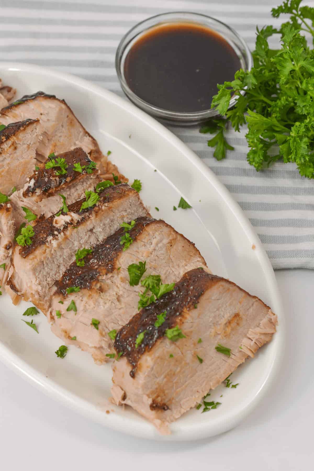 Succulent sliced roast pork tenderloin, slow-cooked to perfection in a crock pot, garnished with parsley on a white plate. It sits beside a small bowl of sauce and a bundle of fresh parsley for an elegant touch.