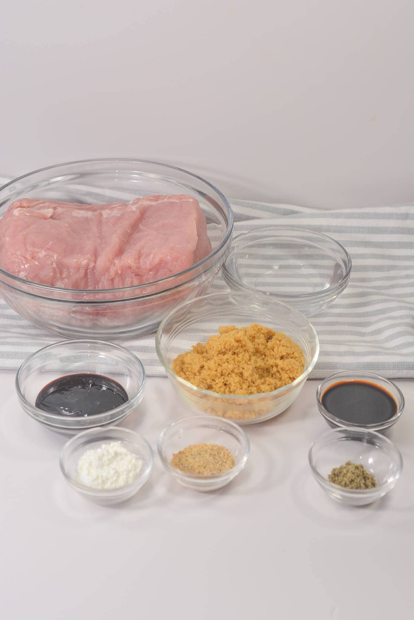Pork tenderloin sits raw in a glass bowl on a striped cloth, surrounded by small bowls of sugar, cornstarch, sauce, and spices—perfect for a crock pot transformation.
