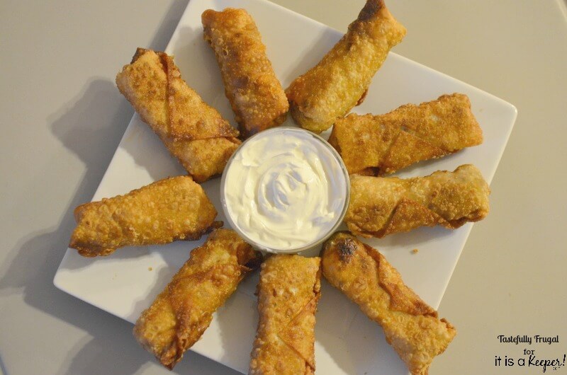 9 Taco Egg Rolls on a white plate with sour cream in a small bowl in the middle. 