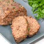A platter garnished with meat with herbs sits on a gray plate. A bunch of fresh parsley is visible in the background.
