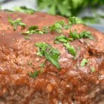 Close-up of a delicious bbq meatloaf topped with parsley, placed on a plate with blurred greenery in the background.