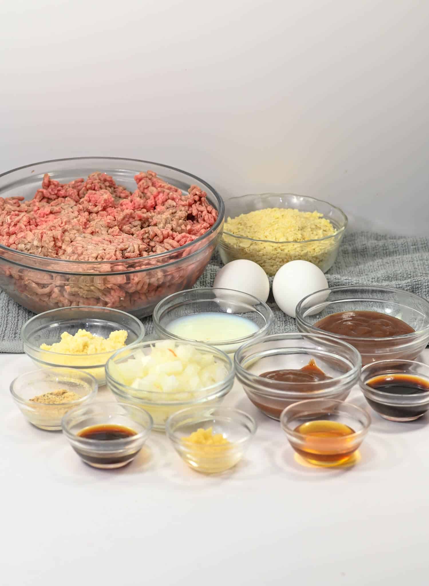 Various ingredients in glass bowls, like those for a BBQ meatloaf, including ground beef, breadcrumbs, eggs, chopped onions, sauces, and seasonings arranged on a surface.