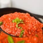 A spoonful of chunky, Hearty Marinara Sauce garnished with fresh basil leaves, held above a pot filled with more savory sauce.