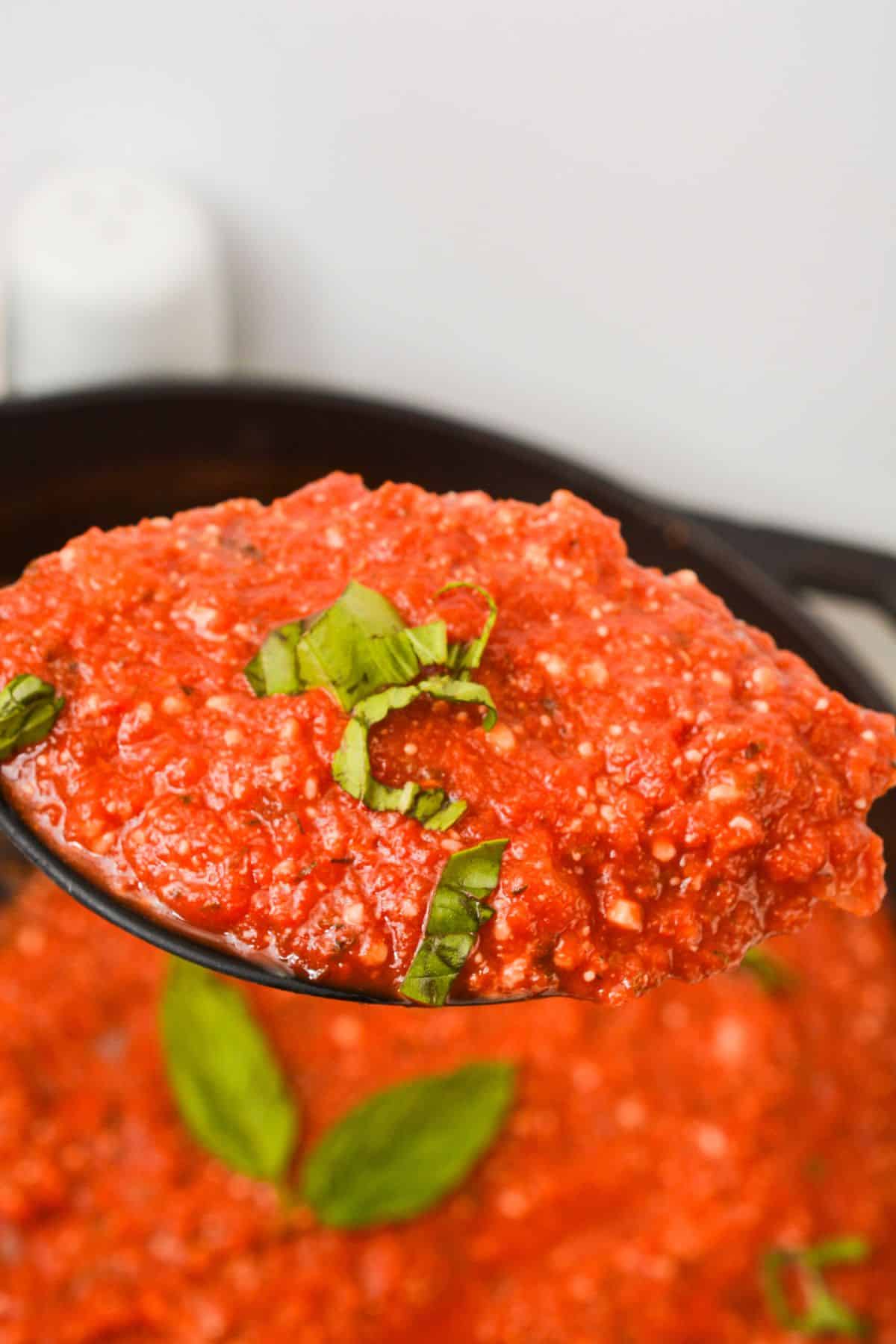 A spoonful of chunky, Hearty Marinara Sauce garnished with fresh basil leaves, held above a pot filled with more savory sauce.