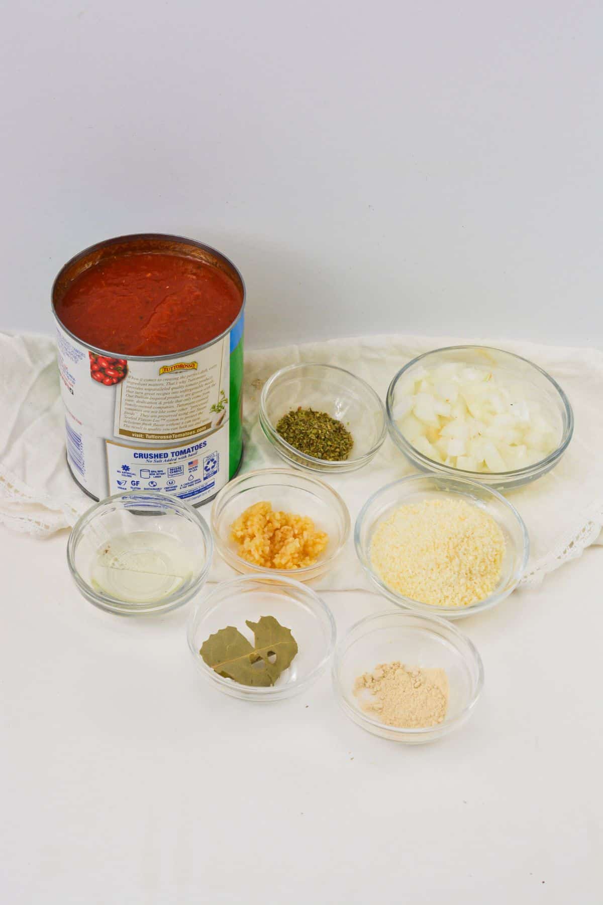 A can of crushed tomatoes and small bowls containing chopped onions, minced garlic, dried oregano, grated cheese, bay leaves, oil, and seasoning.