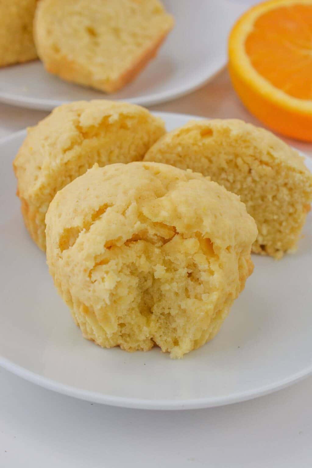 Three Greek Yogurt Muffins, one split in half, rest on a white plate. An orange slice lingers in the background, adding a citrusy flair.