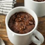 Chocolate chip mug cakes nestled in white mugs sit invitingly on a wooden table, accompanied by a striped cloth and a spoon.