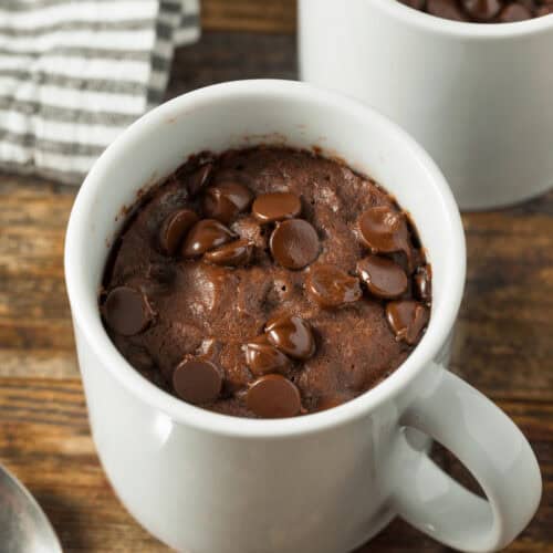 Chocolate chip mug cakes nestled in white mugs sit invitingly on a wooden table, accompanied by a striped cloth and a spoon.