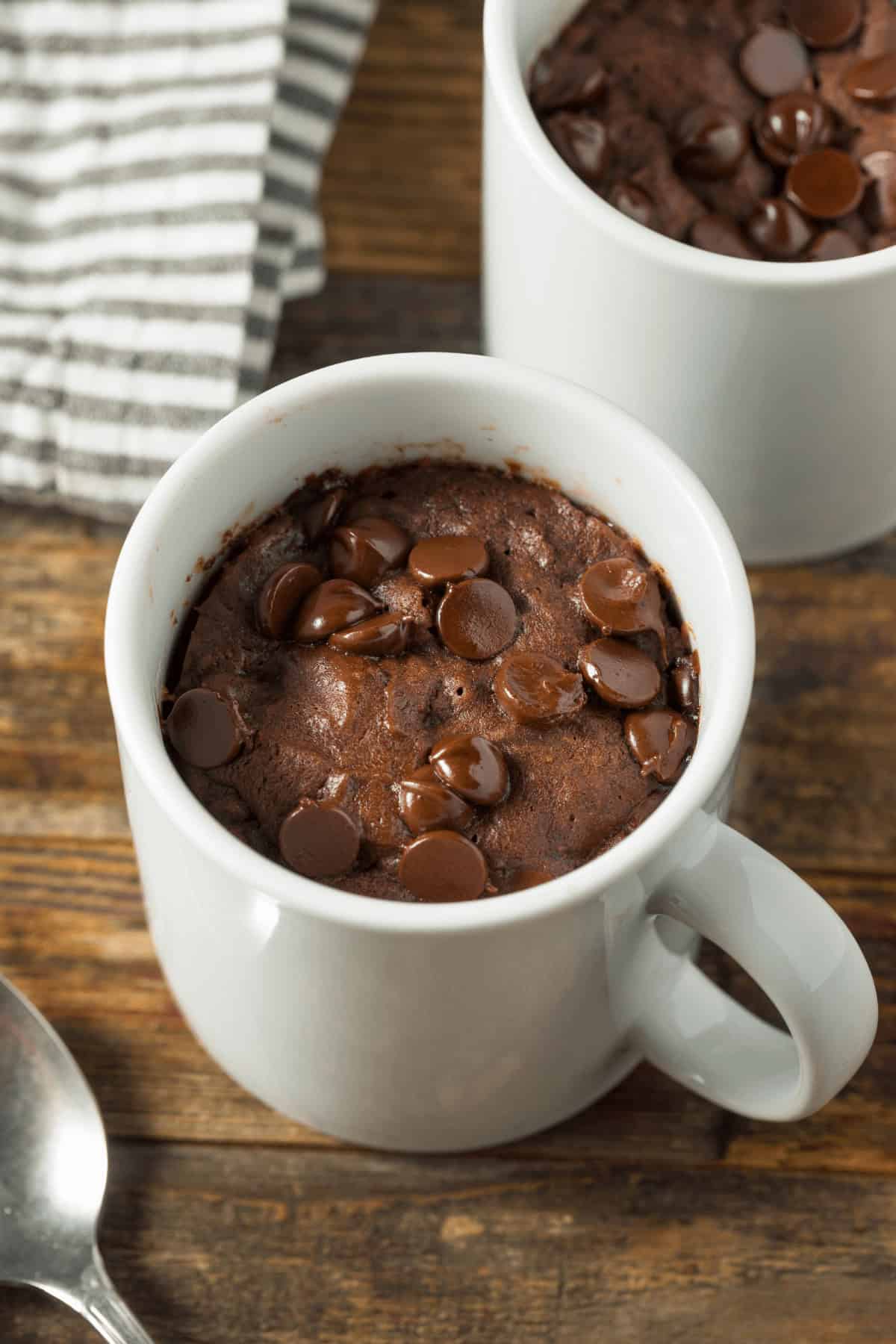 Chocolate chip mug cakes nestled in white mugs sit invitingly on a wooden table, accompanied by a striped cloth and a spoon.