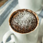 Two white mugs filled with chocolate chip mug cake topped with powdered sugar rest on a striped cloth.