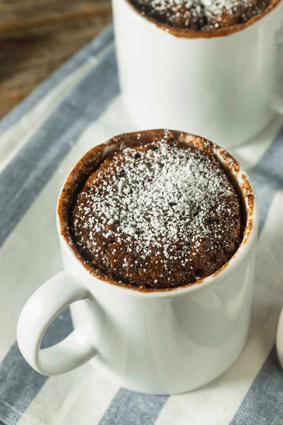 Two white mugs filled with chocolate chip mug cake topped with powdered sugar rest on a striped cloth.