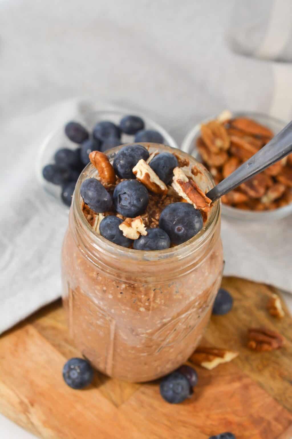 A mason jar filled with tiramisu overnight oats, topped with fresh blueberries and pecans, sits on a wooden surface with bowls of blueberries and pecans in the background.
