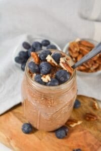 A mason jar filled with tiramisu overnight oats, topped with fresh blueberries and pecans, sits on a wooden surface with bowls of blueberries and pecans in the background.