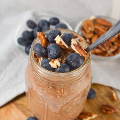 A mason jar filled with tiramisu overnight oats, topped with fresh blueberries and pecans, sits on a wooden surface with bowls of blueberries and pecans in the background.