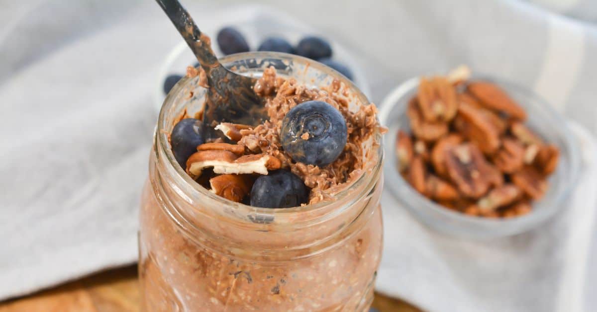 A jar of Tiramisu Overnight Oats topped with fresh blueberries and pecans, with a fork inside. A small bowl of pecans and blueberries is in the background.
