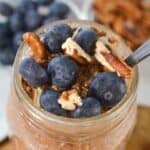 A mason jar filled with tiramisu overnight oats, topped with fresh blueberries and pecan pieces, with a spoon inside.